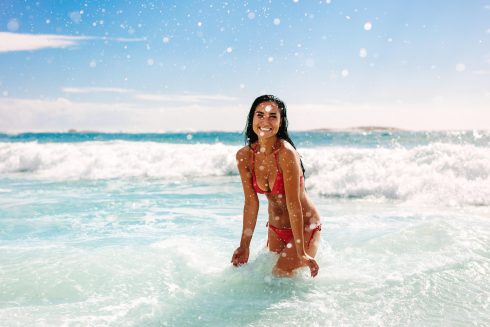 Woman On Vacation Having Fun At The Beach Playing In The Water. Female In Bikini Enjoying Splashing Water At The Beach.